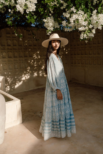 Jasmine Maxi Dress Sea Mist — architectural lifestyle shot under bougainvillea, carved stone wall Jaipur, straw hat, hand block print sea mist blue cotton maxi, artisan ethical slow fashion | Daughters of India