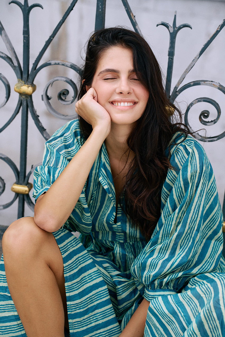 Smiling woman seated against ornate metal gates wearing the Daughters of India Kyra Midi Dress in Spearmint with bare legs showing the midi length