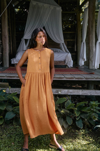 Nadi Dress Amber — full length front view in tropical garden, amber double gauze cotton midi dress with wooden button front and gathered waist, hands in pockets | Daughters of India